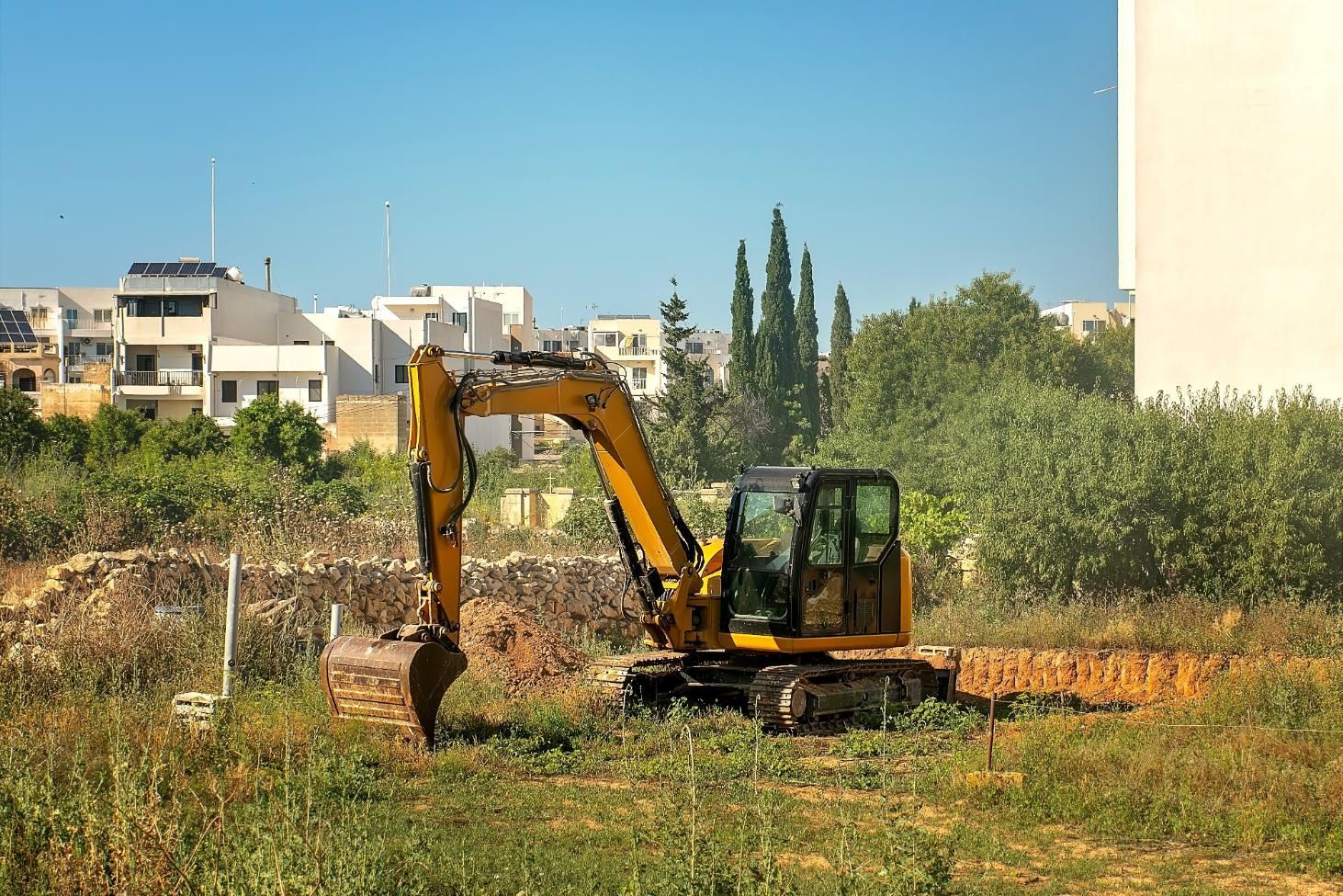 Yellow excavator working in a grassy field with buildings and trees in the background