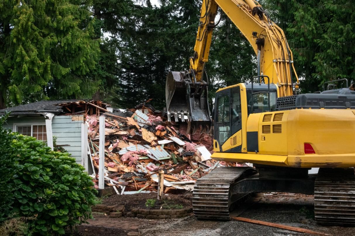 Yellow excavator demolishing a small house amid rubble and trees