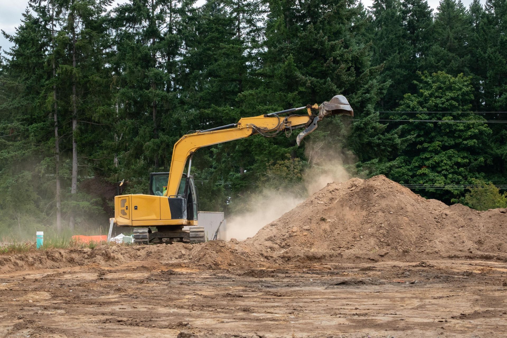 Yellow excavator dumping soil onto a dirt pile at a construction site with trees in the background