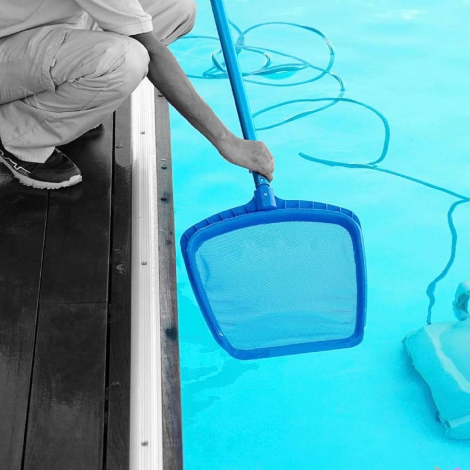 A person crouches on a wooden deck, using a blue mesh pool skimmer to clean a bright blue swimming pool.