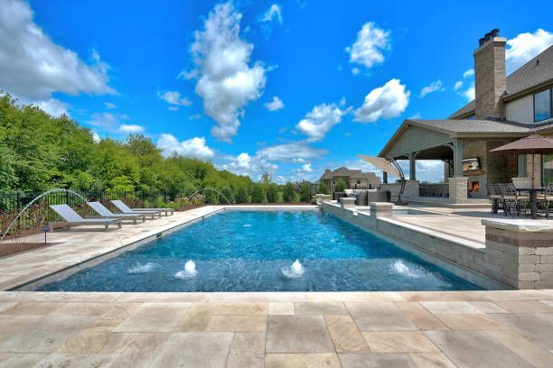 A rectangular swimming pool with fountains and lounge chairs on a stone patio next to a luxury home under a blue sky.