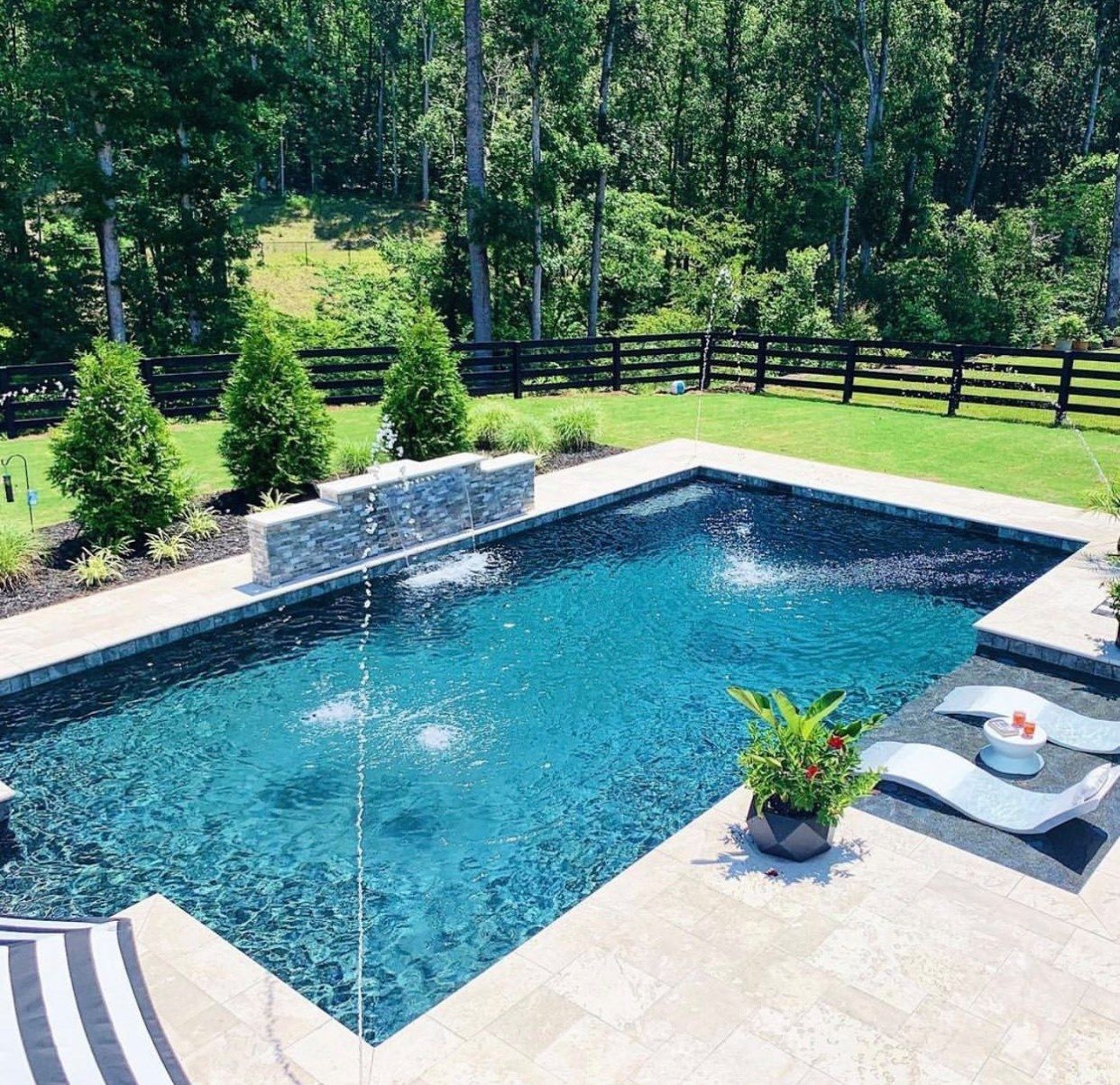 A person in blue cleaning a turquoise swimming pool with a long-handled net and vacuum hose from a wooden deck.