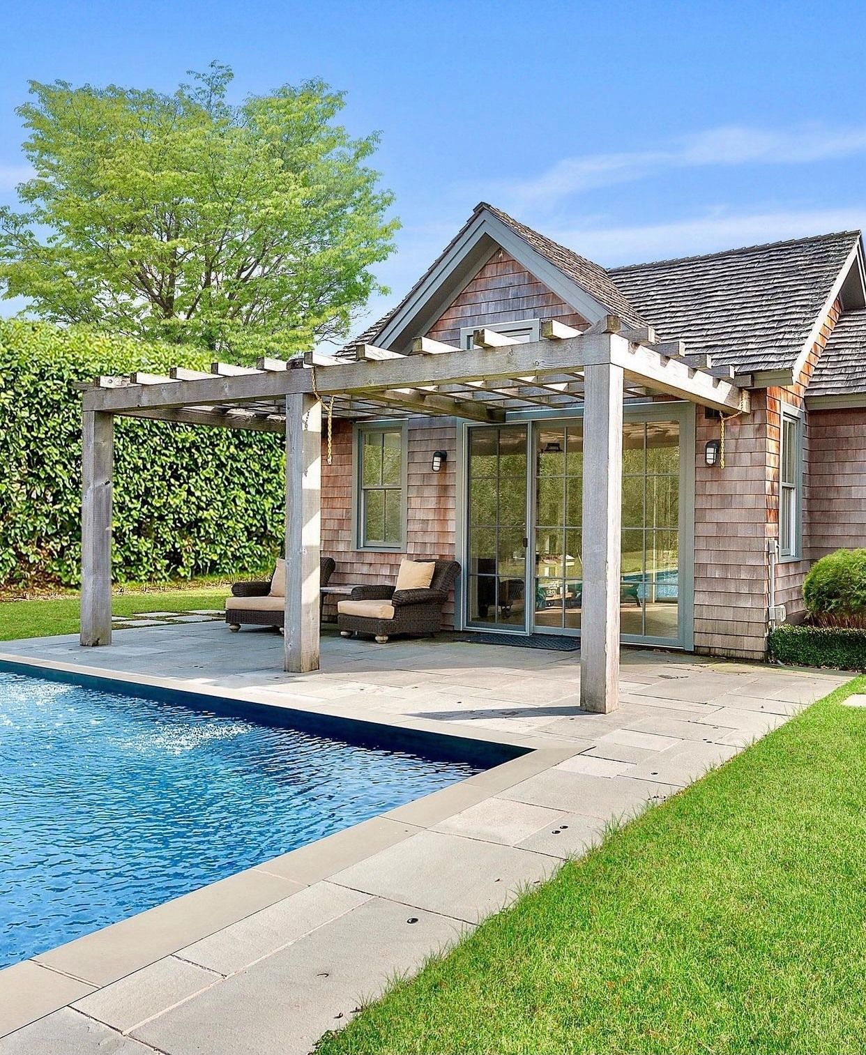 A pool next to a brick patio with a wooden pergola, outdoor seating, and a house exterior under a blue sky.