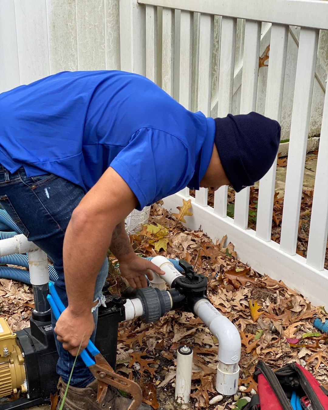A person in a blue shirt and beanie uses pliers to work on outdoor plumbing pipes next to a white fence and fallen leaves.