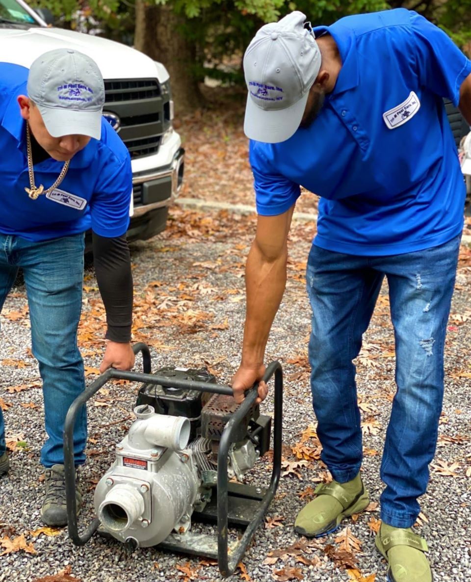 Two people in matching blue shirts and grey hats carry a portable water pump outdoors near a white vehicle.