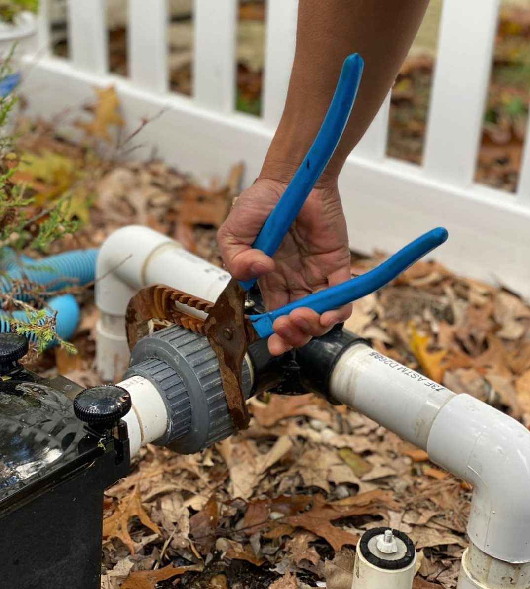A person uses blue-handled pliers to turn a rusty metal fitting on white PVC pipes in an outdoor, leaf-covered setting.