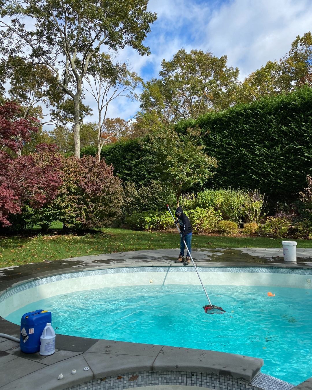 A person cleans a round swimming pool with a long-handled net in a sunny, tree-filled backyard.