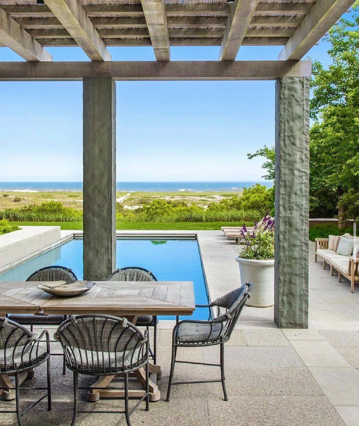 A stone-pillared patio with a dining set and pool overlooking a grassy landscape and the ocean on a clear day.