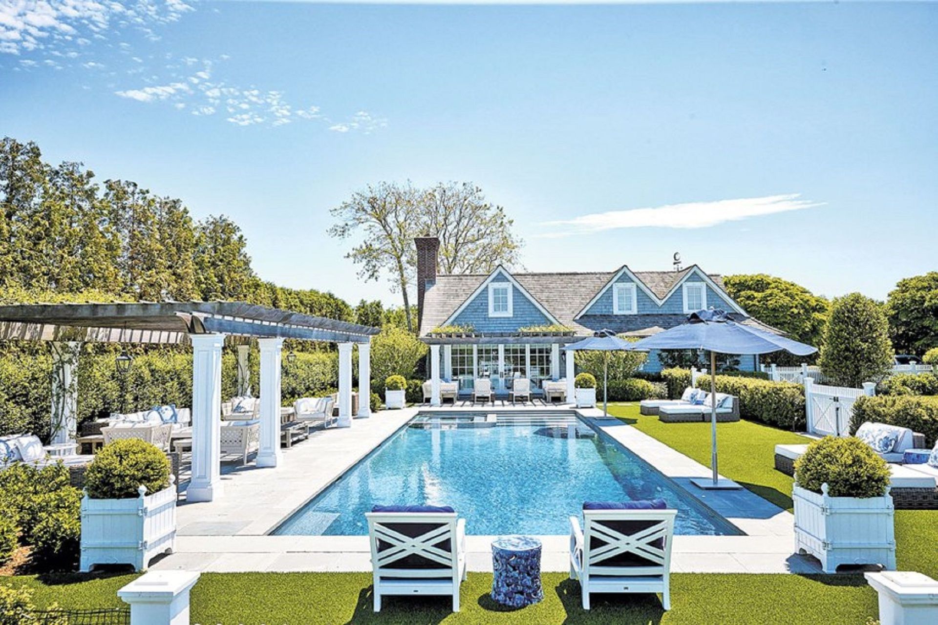 A sunny backyard featuring a rectangular swimming pool, outdoor seating, a pergola, and a cottage with blue shingles.
