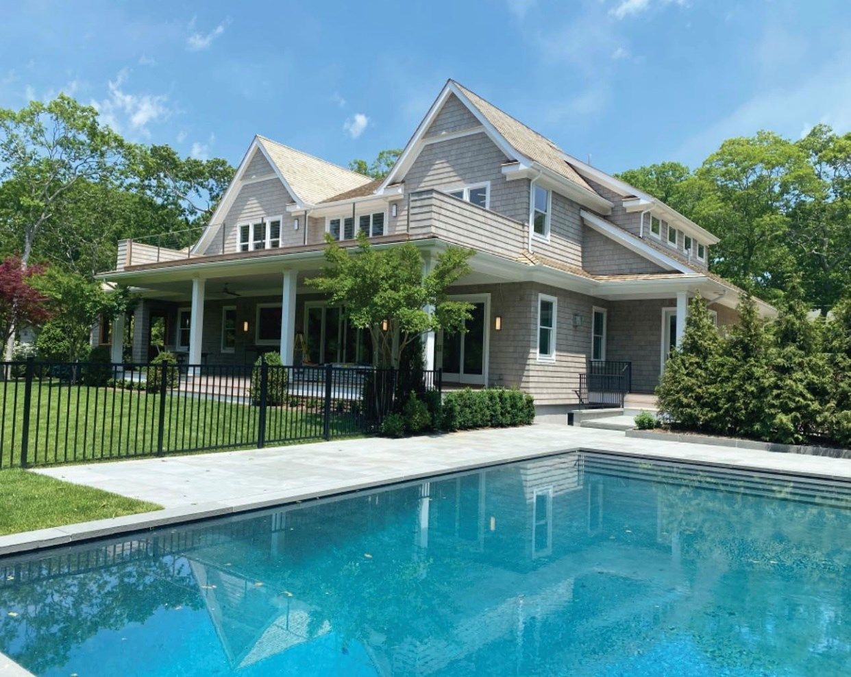 A two-story shingled house with a covered porch overlooking a backyard swimming pool under a blue sky.