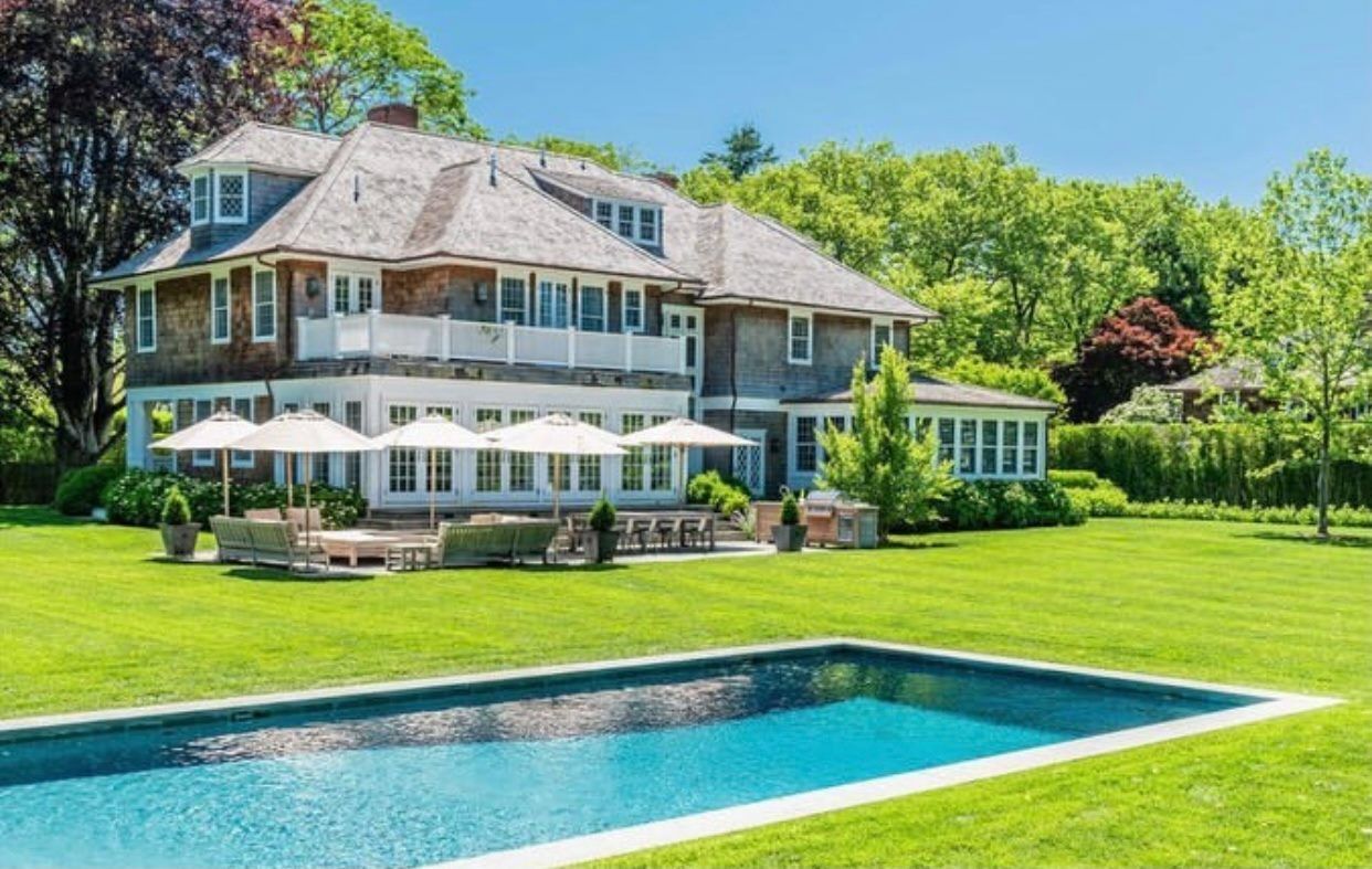 A large shingled house with a white terrace, patio umbrellas, and a rectangular swimming pool on a lush green lawn.