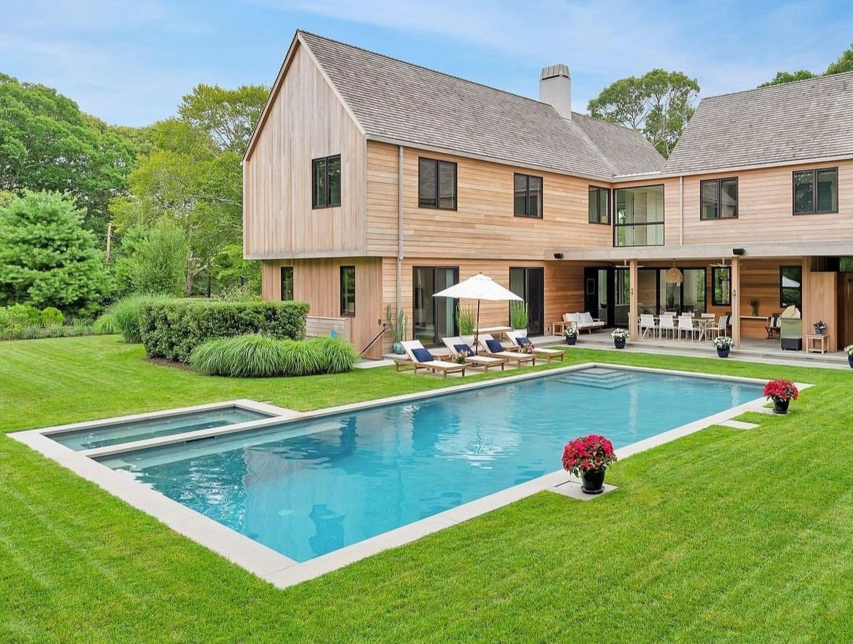 A rectangular swimming pool with a spa on a green lawn in front of a modern, two-story wooden house.