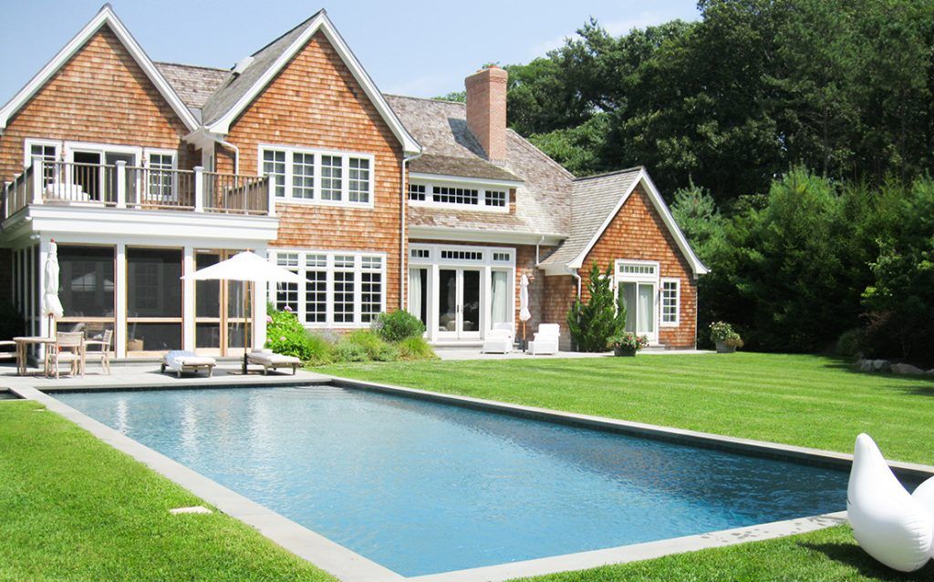 A large house with shingled siding, a screen porch, and a rectangular swimming pool set in a spacious green lawn.