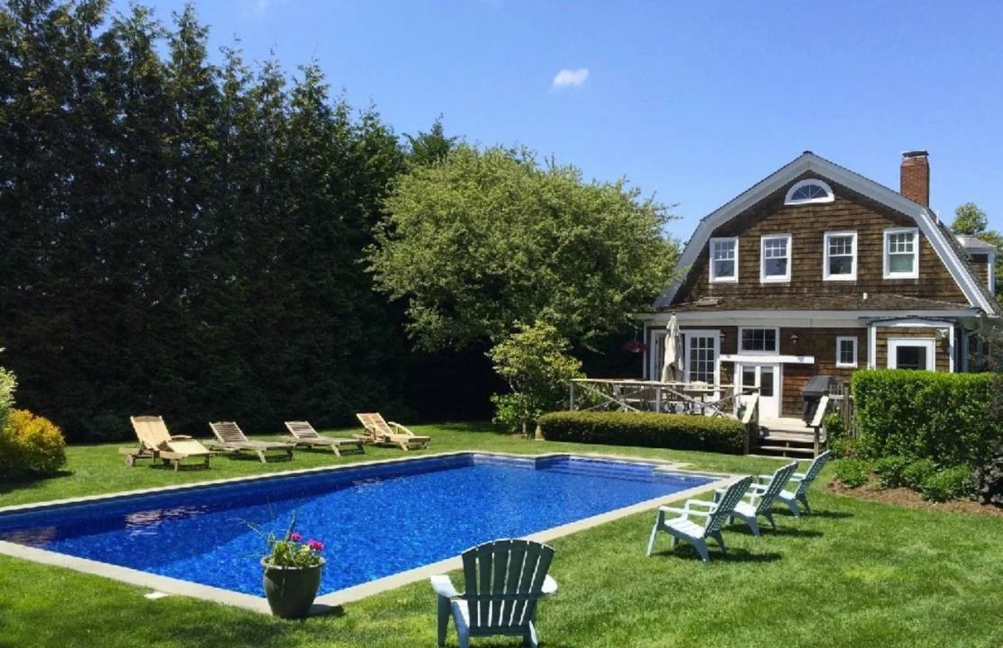 A rectangular swimming pool with blue water sits in a green lawn next to a brown, cedar-shingled house on a sunny day.
