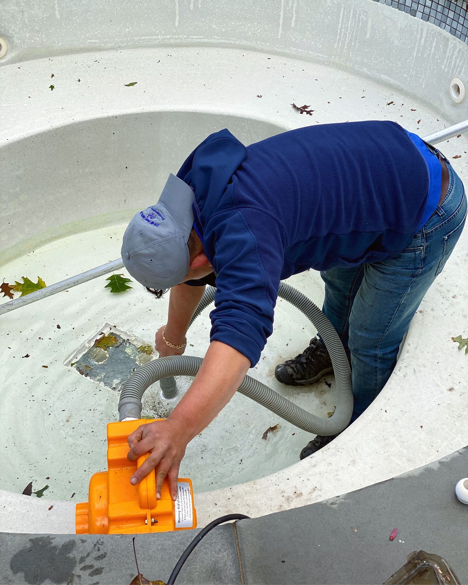 A person in a blue hoodie and gray cap cleans a pool with a bright orange vacuum tool.