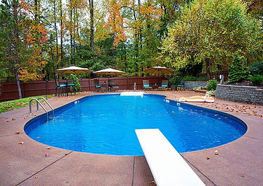 A swimming pool surrounded by a patio and autumn trees in a backyard setting, featuring a diving board in the foreground.