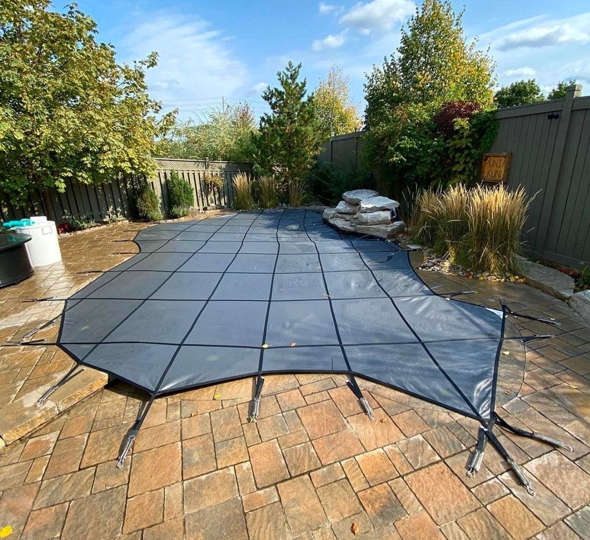 A gray, grid-patterned safety cover stretched over a kidney-shaped swimming pool on a paved patio in a fenced backyard.