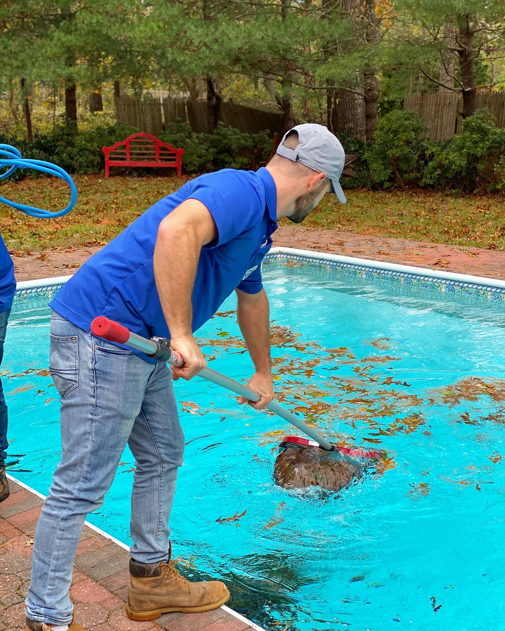 A person in a blue shirt and jeans stands by a backyard pool, using a pole net to skim fallen leaves from the water.