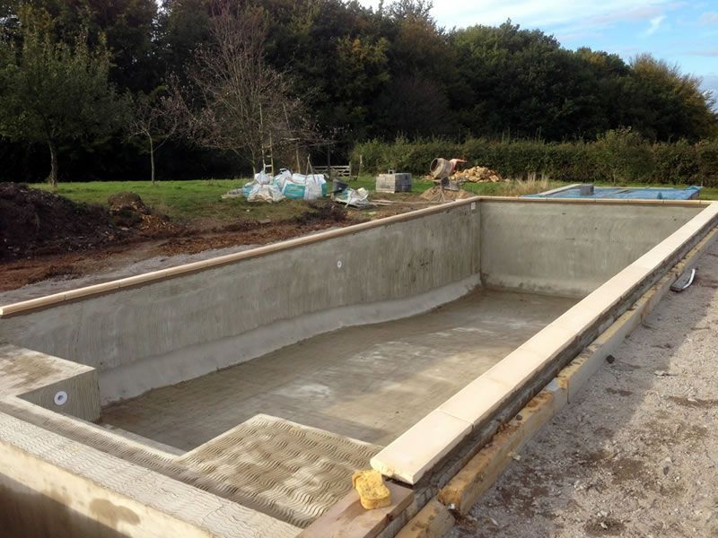 An unfinished concrete swimming pool in a backyard construction site with a light-colored stone coping.