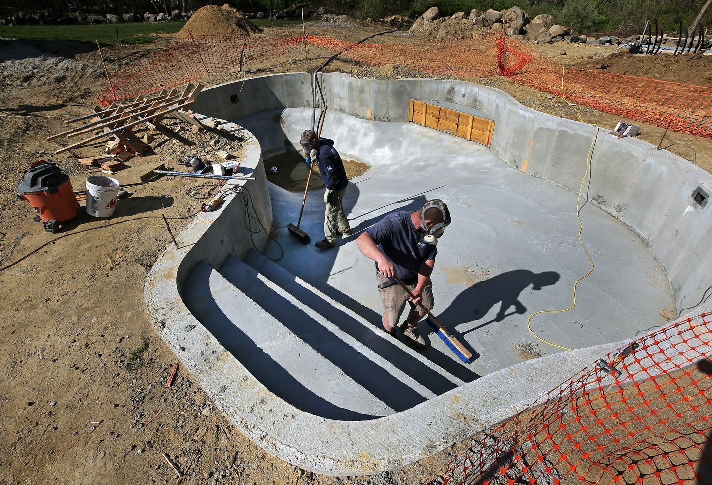 Two workers clean and prepare the surface of a concrete swimming pool under construction outdoors.