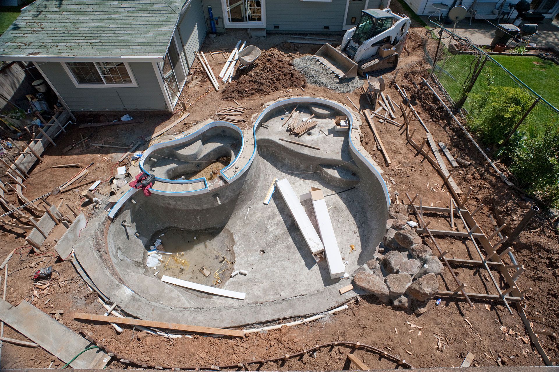 High-angle view of a backyard undergoing pool construction, showing a concrete pool shell, excavation, and equipment.