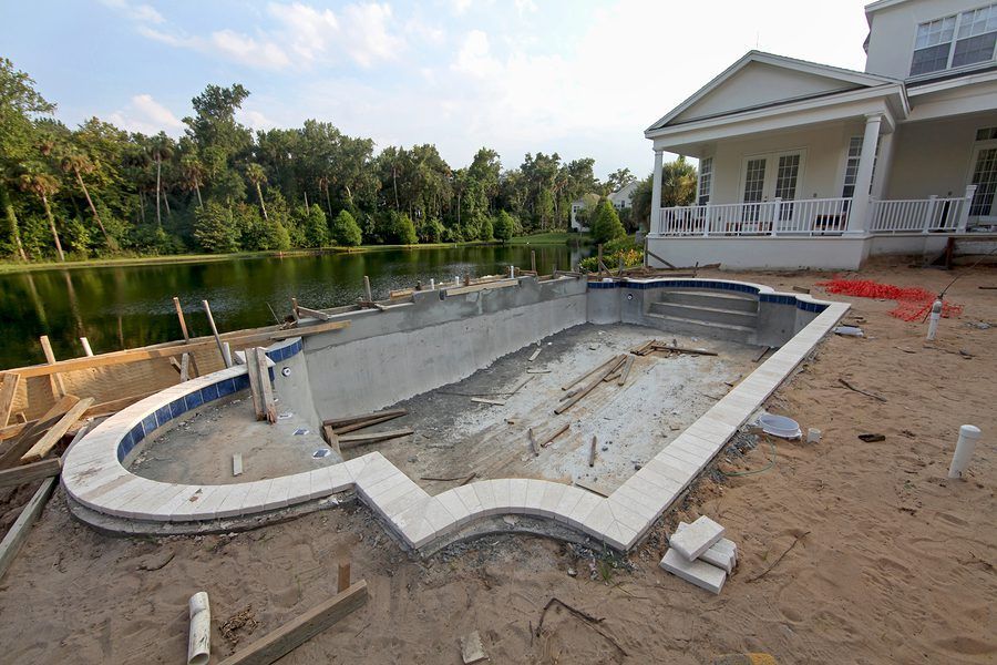 An unfinished rectangular swimming pool with curved edges, under construction next to a house near a lake.