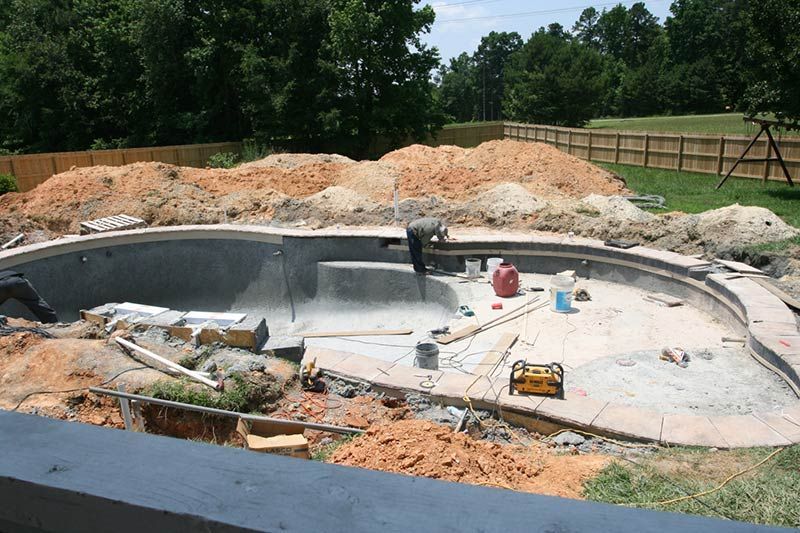 A swimming pool under construction in a backyard, featuring concrete, excavation piles, and a worker.