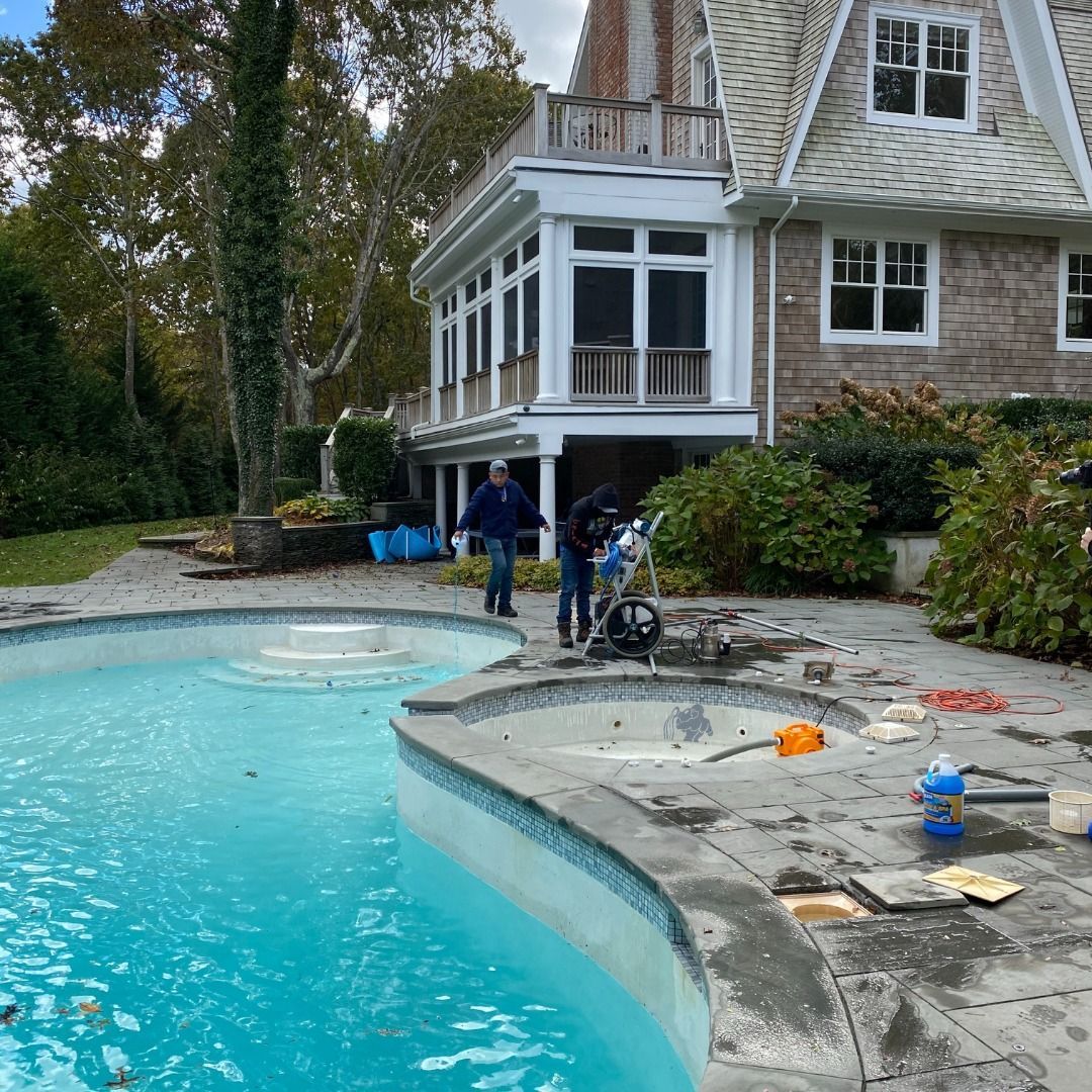 Two people work on a pool deck near a swimming pool and spa, next to a large house with a porch and cedar shingle siding.