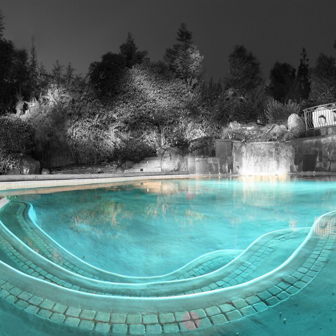 Curved stone steps lead into a bright turquoise swimming pool at night, set against a black and white wooded backdrop.