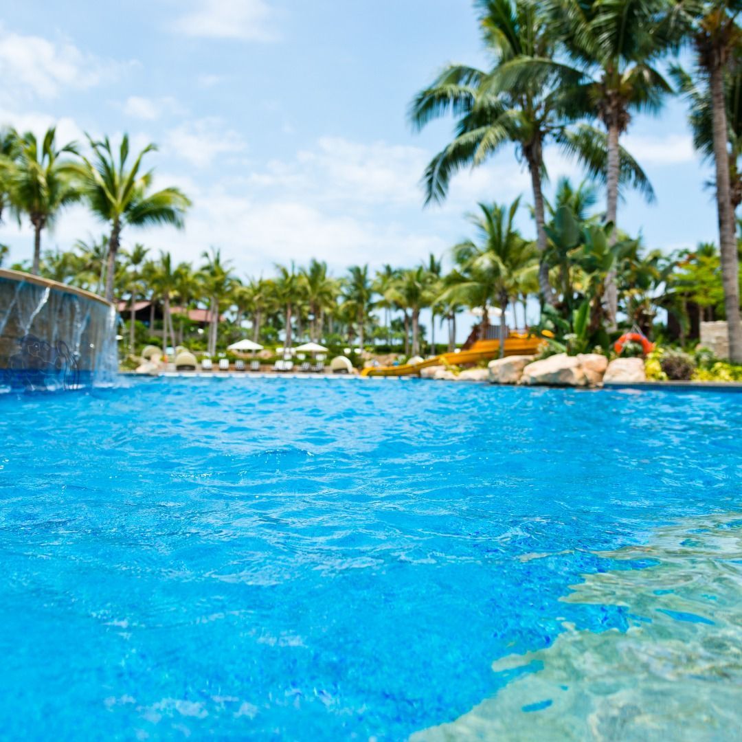 A bright blue swimming pool with a stone waterfall feature, surrounded by lush palm trees under a sunny sky.