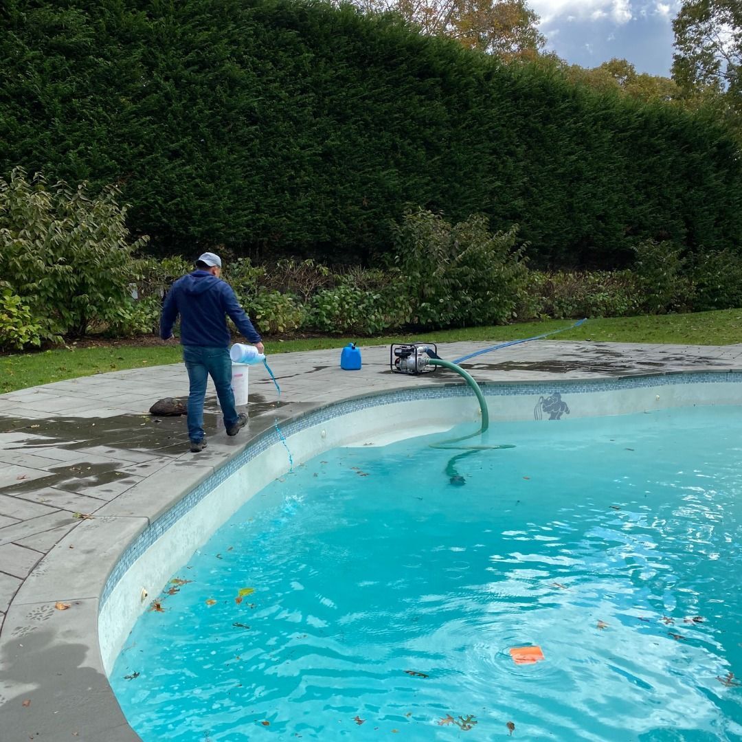 A person in a blue jacket walks on a pool deck next to a swimming pool being cleaned with a hose and pump.