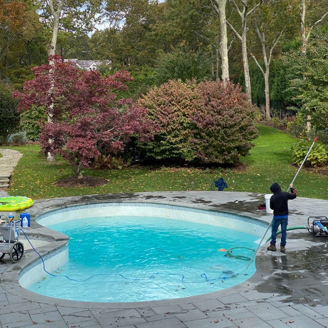 A person cleans a curved backyard swimming pool with a long-handled brush on a stone patio surrounded by trees.