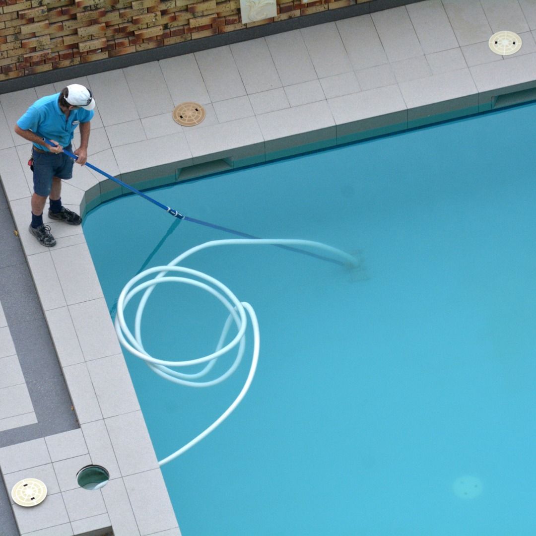 A pool technician in a blue shirt uses a long pole and hose to clean the corner of a light blue swimming pool.