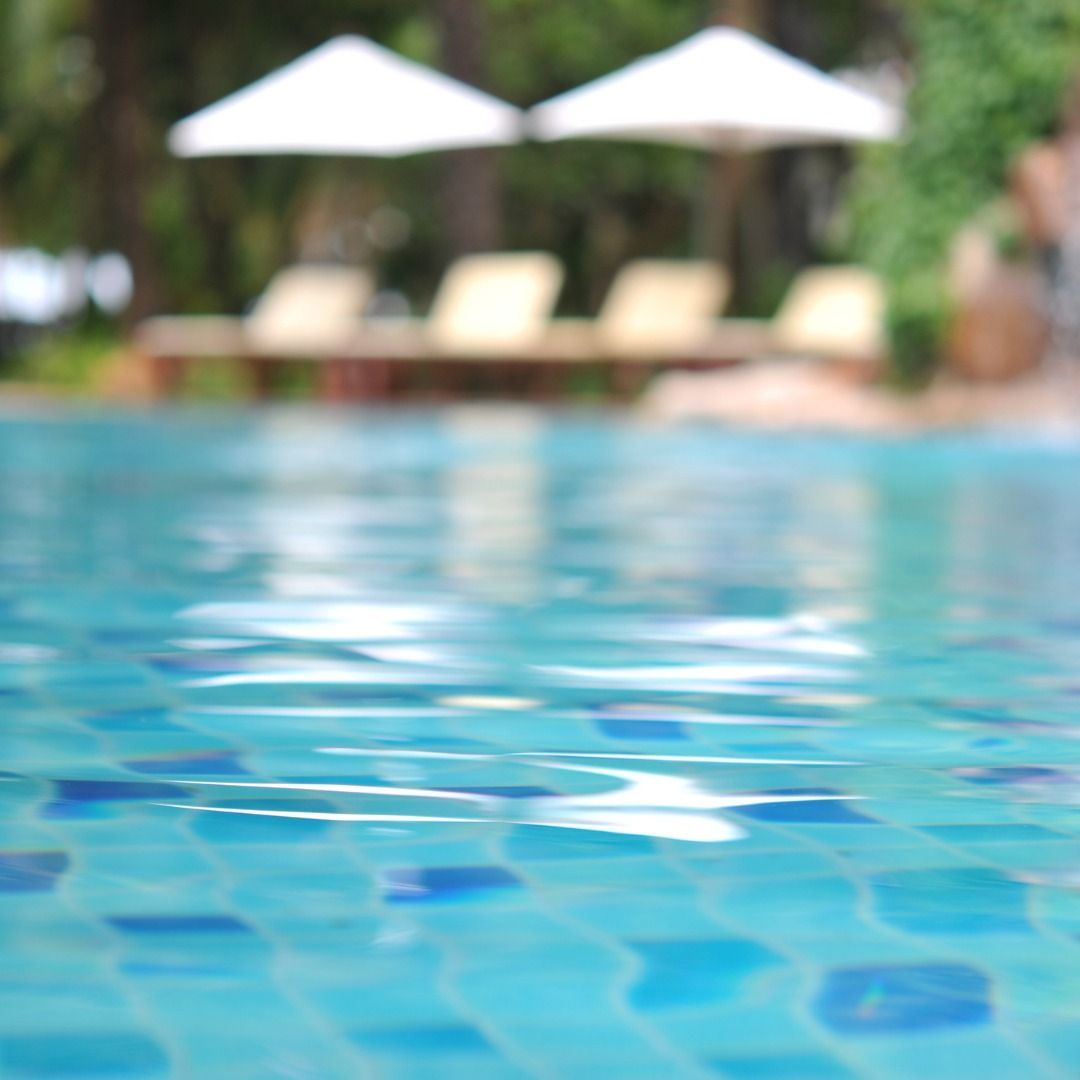 A close-up view of rippling blue pool water with blurred lounge chairs and white umbrellas in the background.