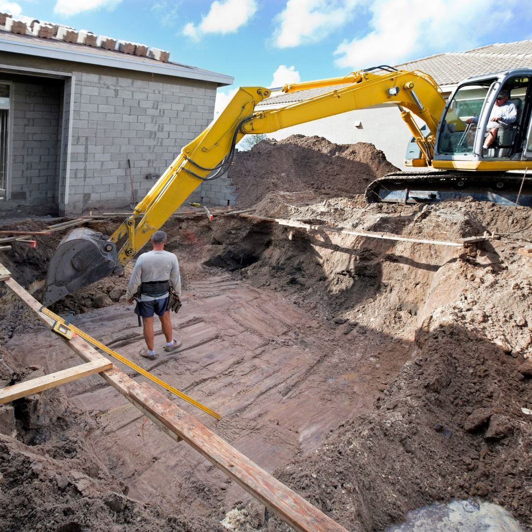 A person in a hard hat stands inside a large excavation pit next to a yellow excavator under a bright blue sky.