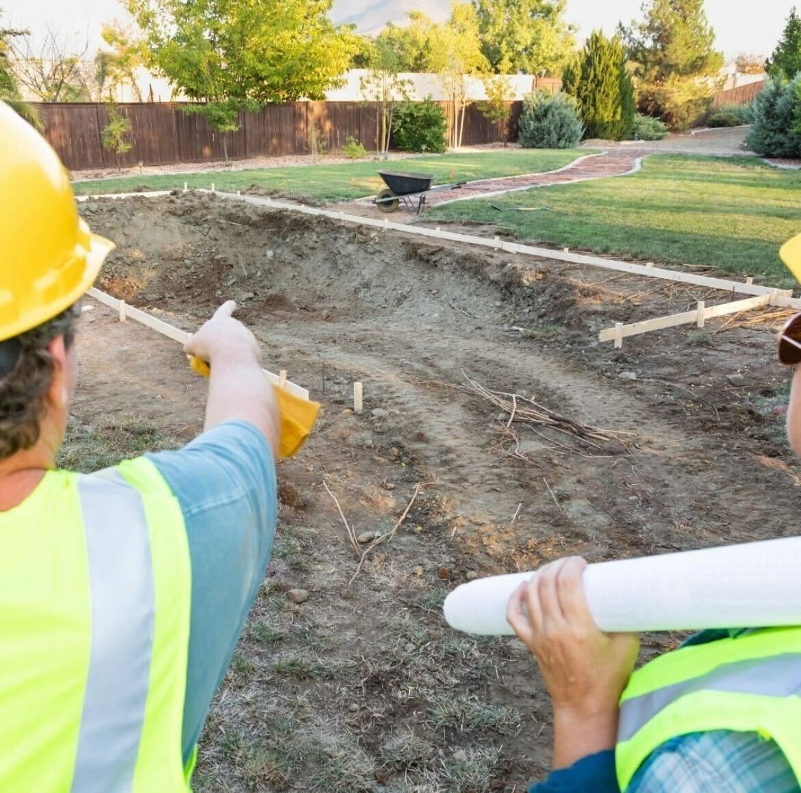 Two construction workers in high-visibility vests and hard hats look over a dirt construction site.