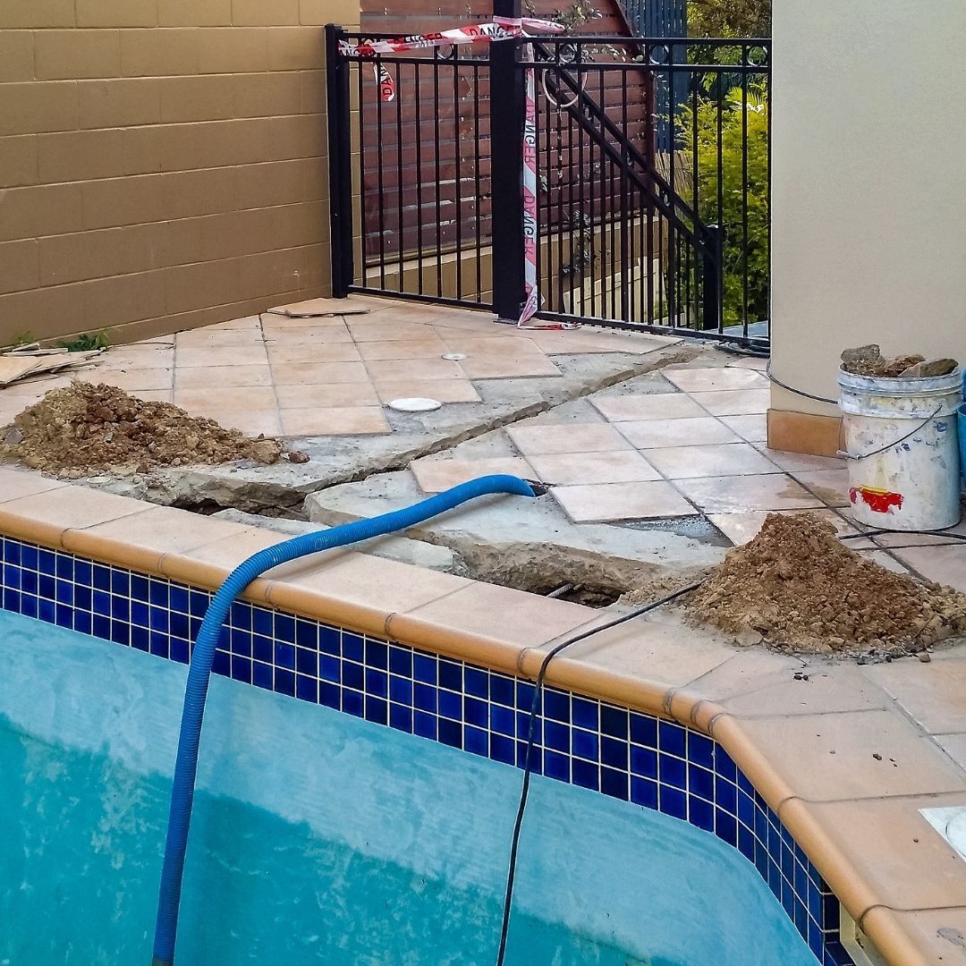 Construction work on a swimming pool deck with missing tiles, loose dirt piles, and a blue hose in the water.