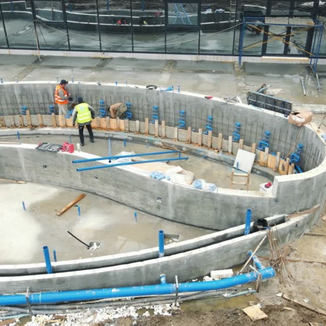 Two workers in safety vests install plumbing pipes inside the curved concrete walls of an unfinished swimming pool.