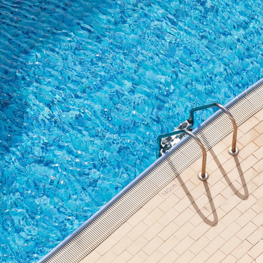 A high-angle view of a swimming pool edge with a metal ladder and light-colored tile decking beside bright blue water.