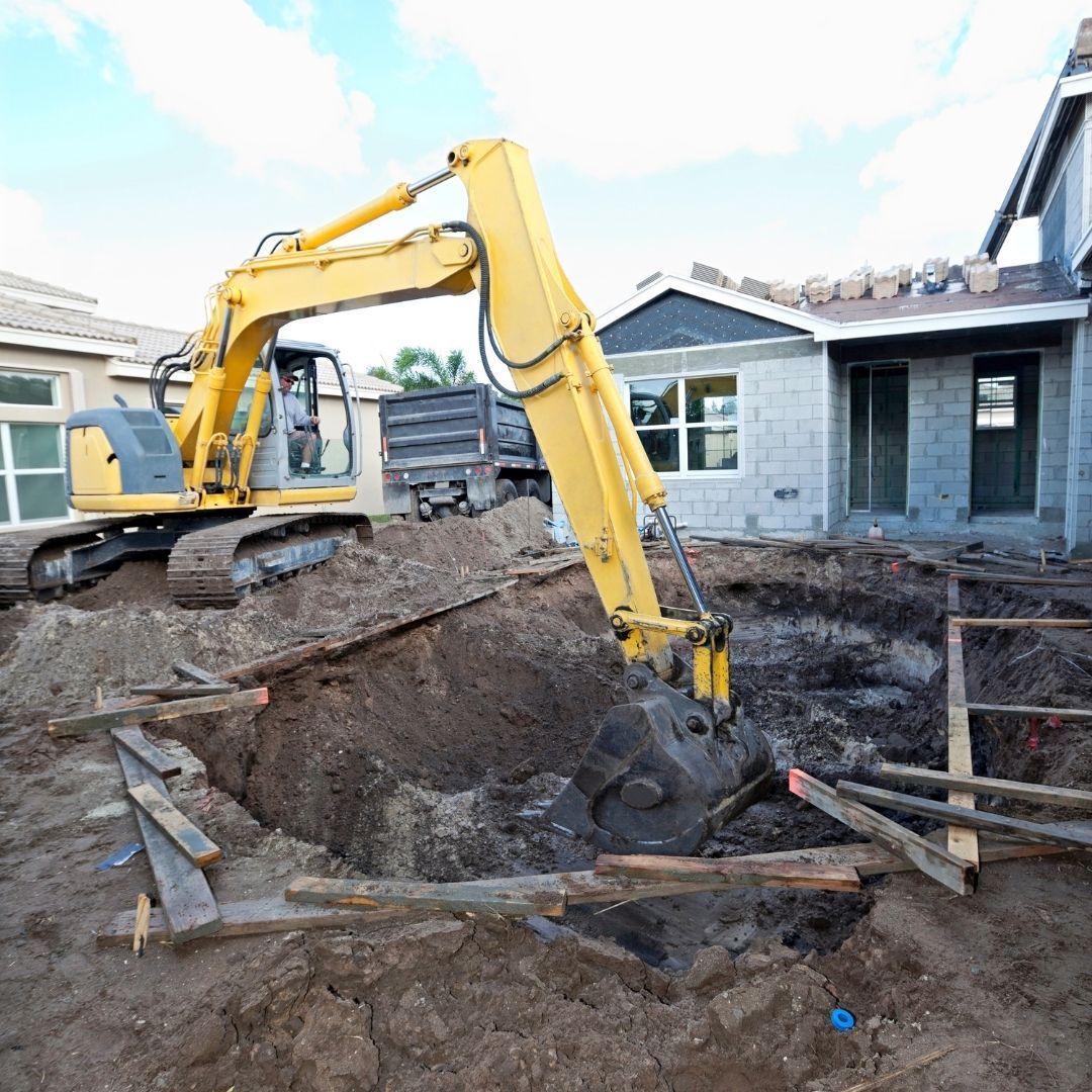A yellow excavator digs a large hole for a backyard pool next to a house under construction.