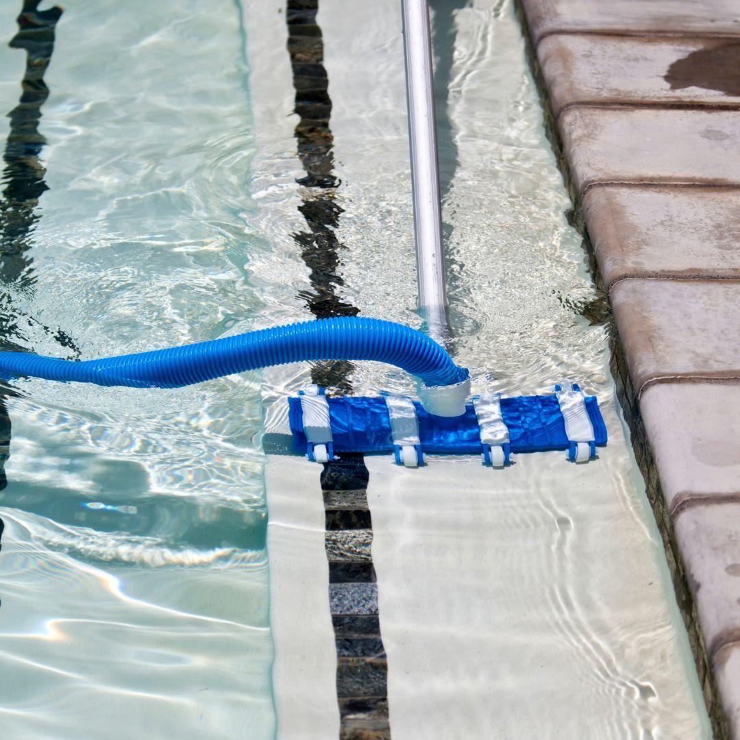 A blue pool vacuum head with a long handle and blue hose cleaning the bottom of a pool alongside the tiled edge.