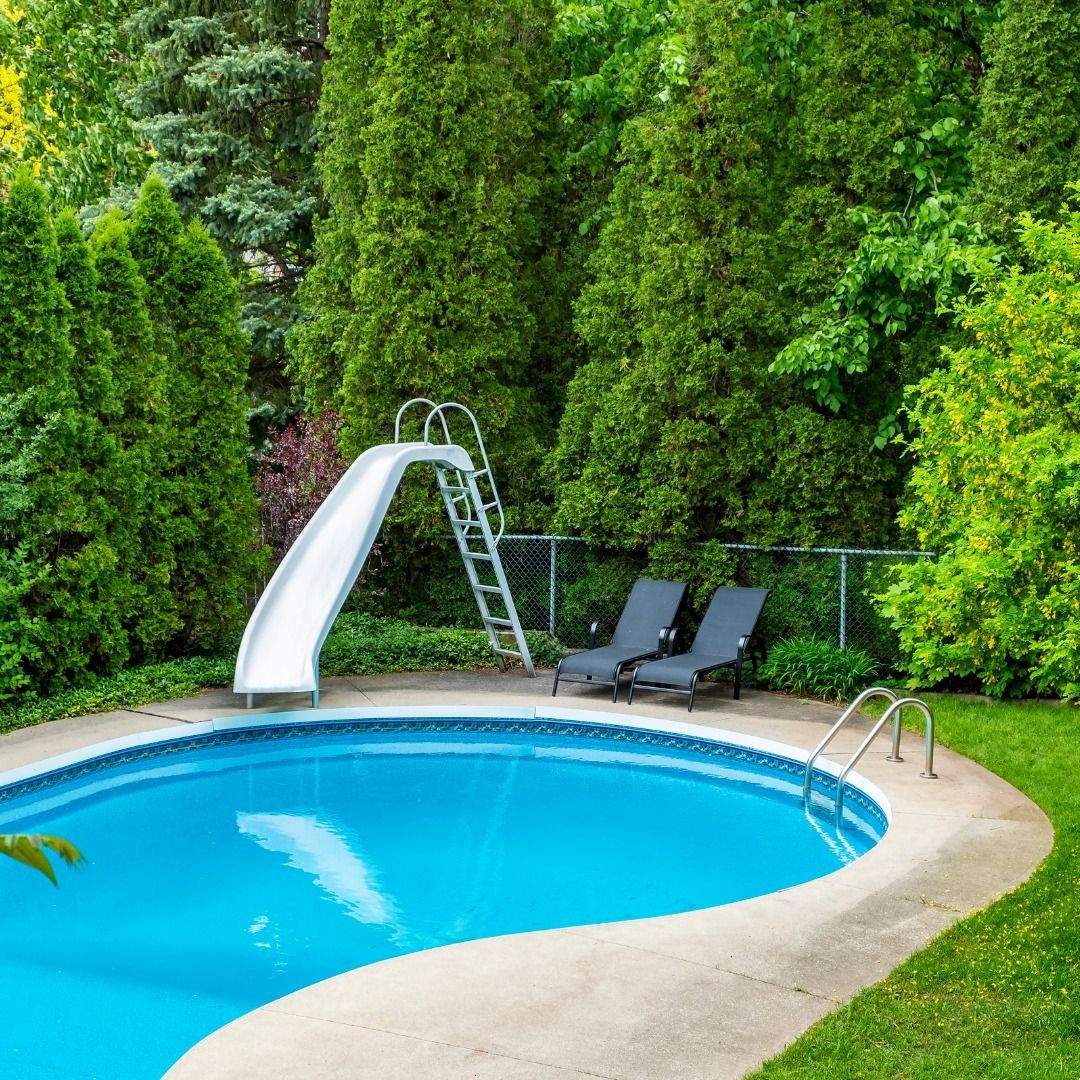 A backyard swimming pool with a curved white slide and two empty lounge chairs on a paved deck surrounded by tall trees.