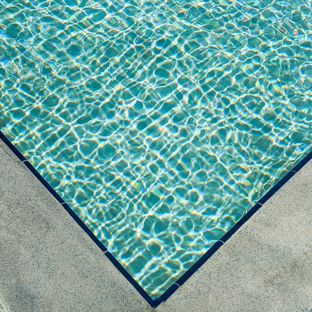 A corner view of a swimming pool with shimmering turquoise water framed by a dark edge and light gray concrete decking.