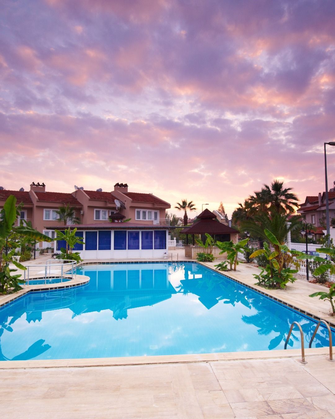 A serene, bright blue swimming pool sits in front of a tan residential building under a vibrant, purple sunset sky.