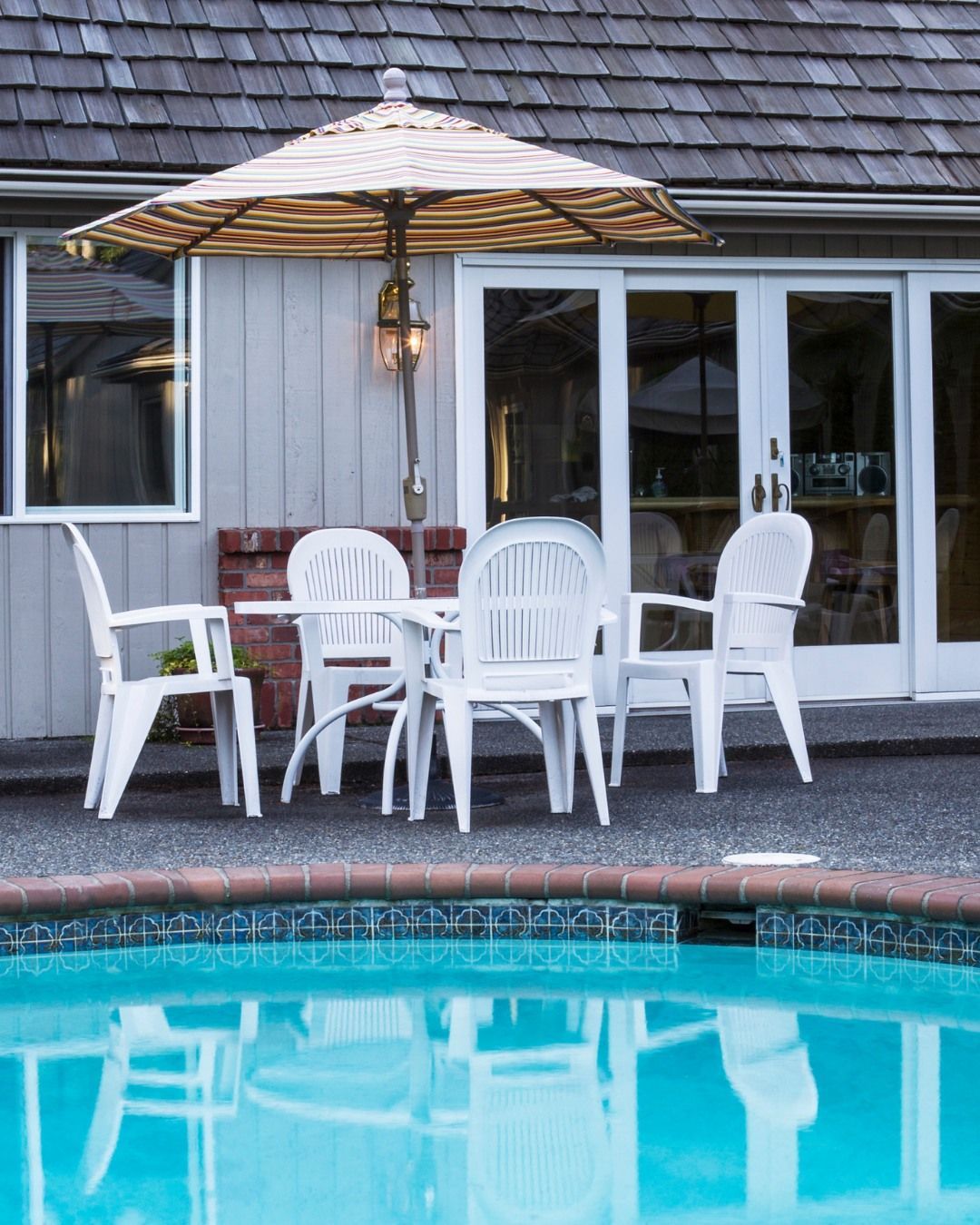 A patio set with white plastic chairs and a table under a striped umbrella sits next to a pool by a building.