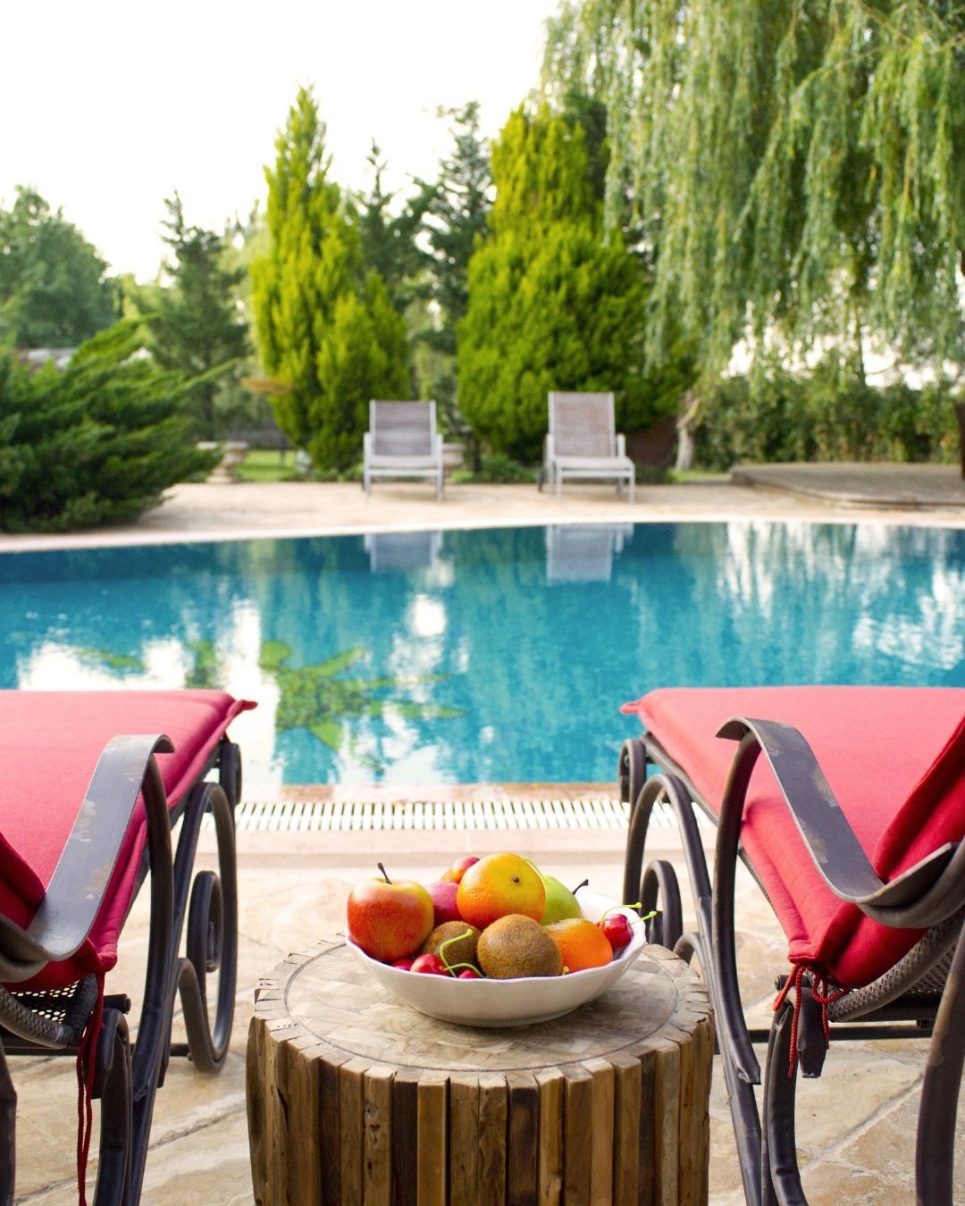 Two red lounge chairs face a swimming pool with a bowl of fruit on a wooden stump table in the foreground.