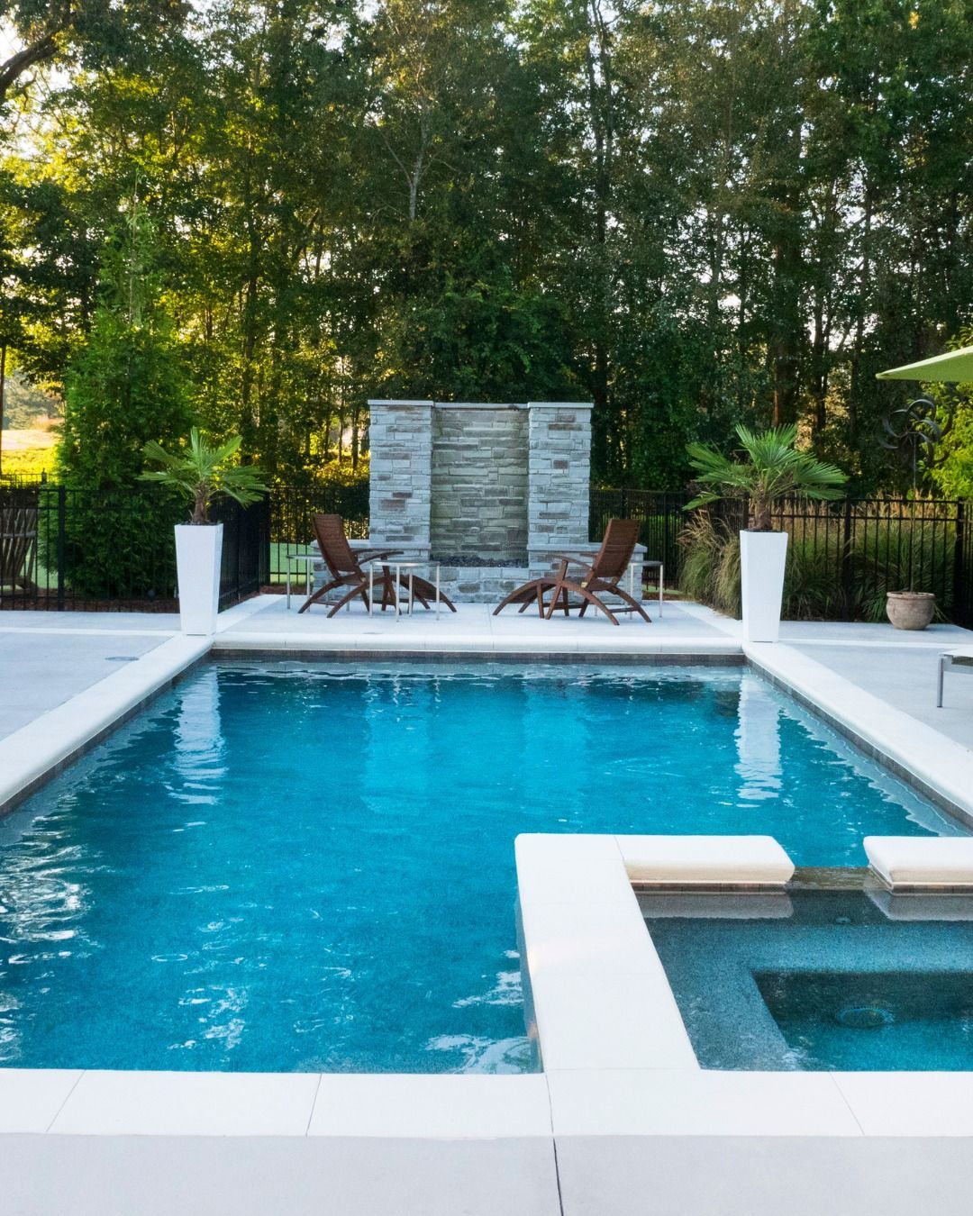 A rectangular swimming pool with a spa, a stone water feature, and lounge chairs on a patio surrounded by trees.
