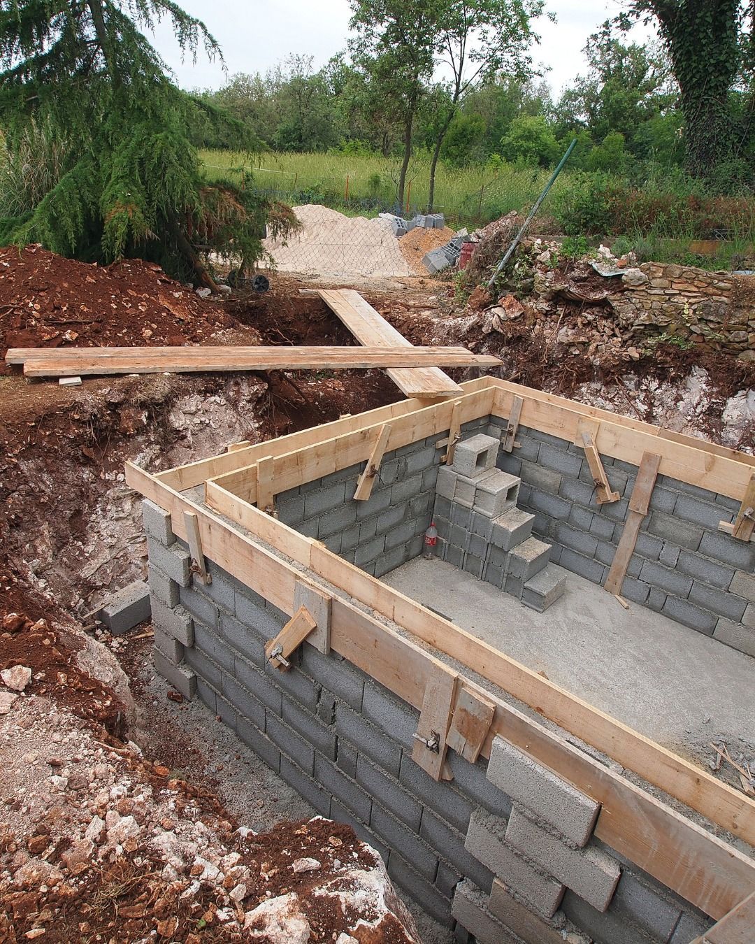 A partially constructed rectangular concrete block foundation in an excavated outdoor site with wooden formwork on top.