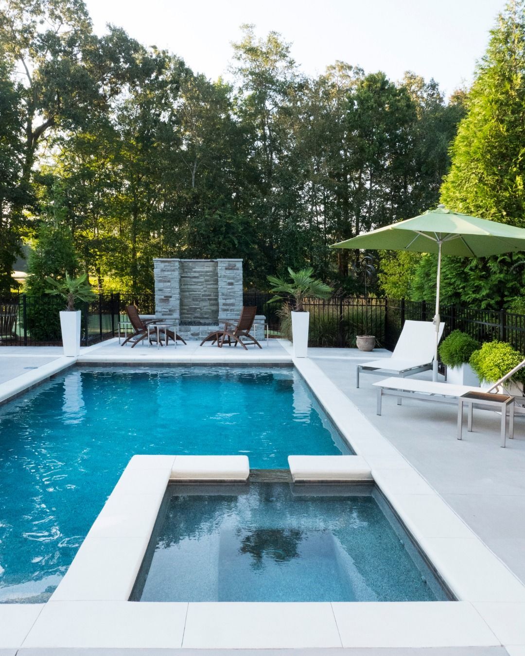 A blue backyard swimming pool with an attached spa, white stone coping, lounge chairs, a green umbrella, and a water wall.