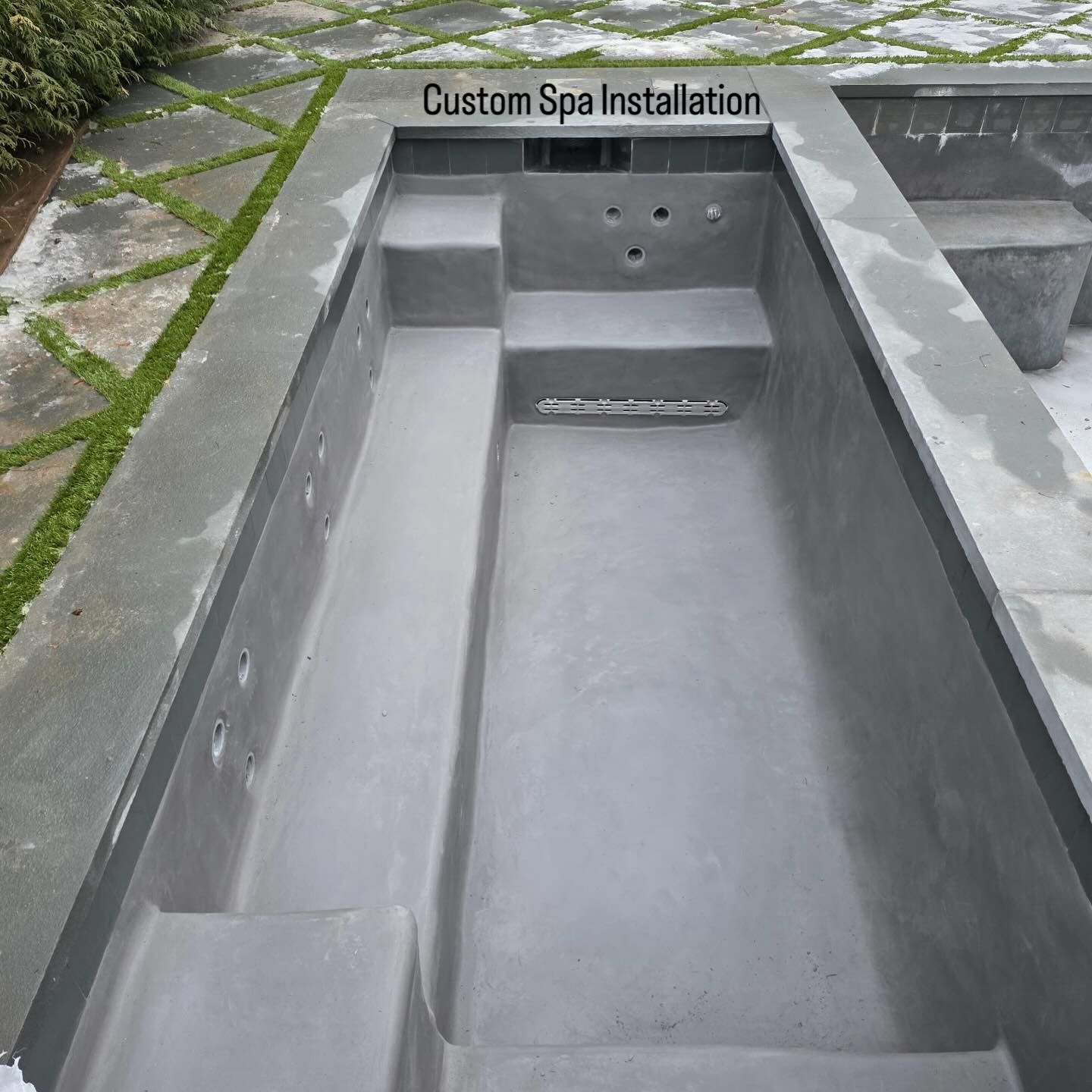 A rectangular gray concrete spa basin under construction, featuring built-in bench seating and jet placements.